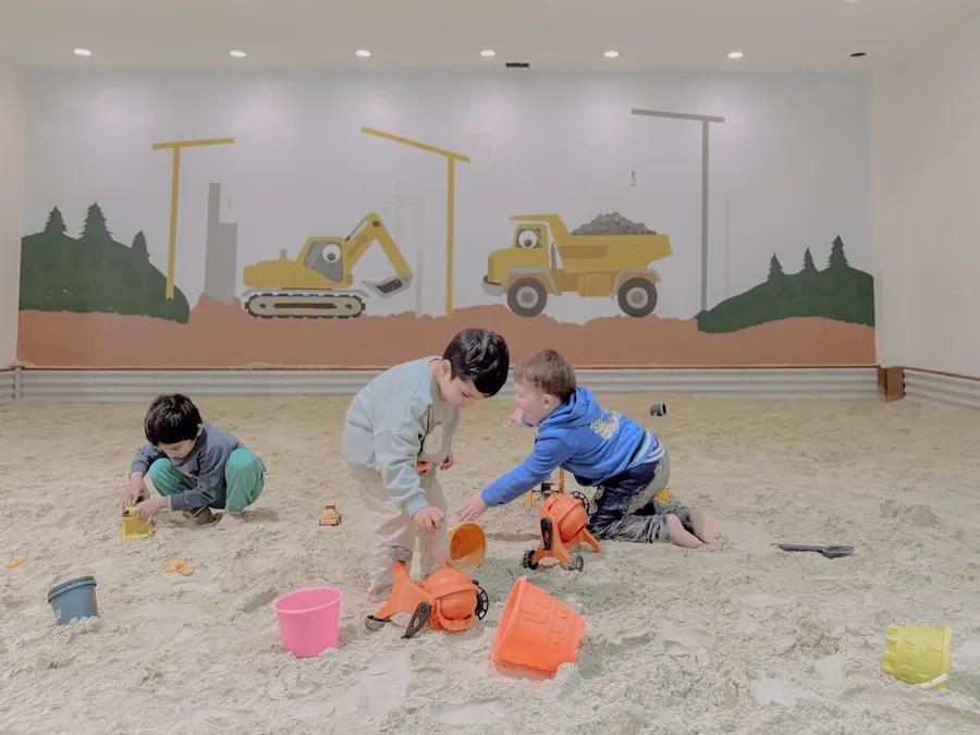 Three children play in an indoor construction-themed sandpit, one tipping sand from a toy mixer.