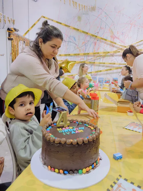 A mother leans in to light the candles on a birthday cake while her son watches in a yellow party hat.