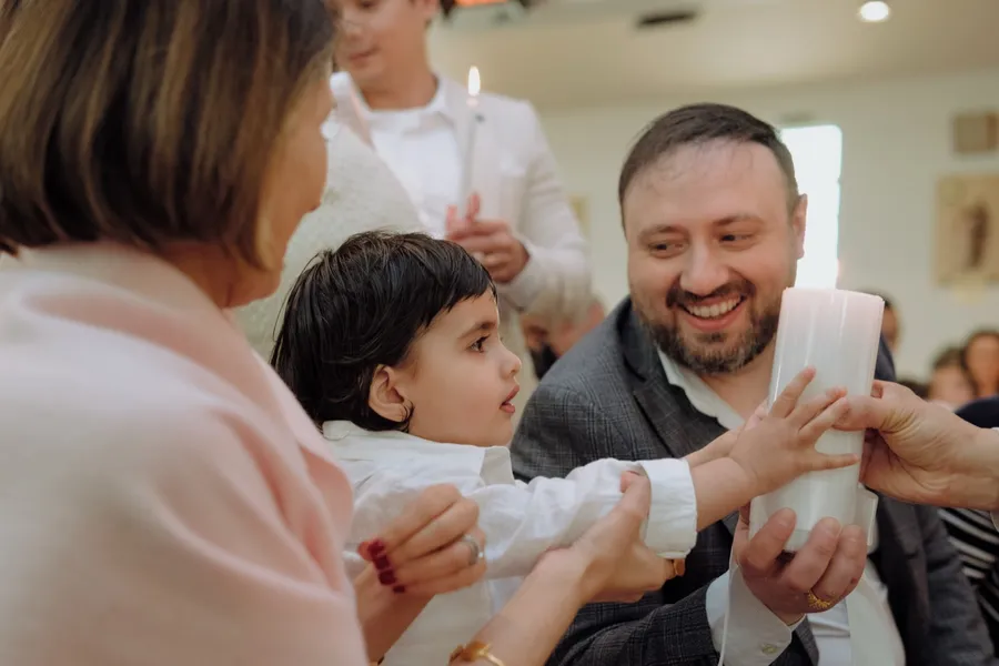 A toddler reaches for a white christening candle as his family smiles around him.