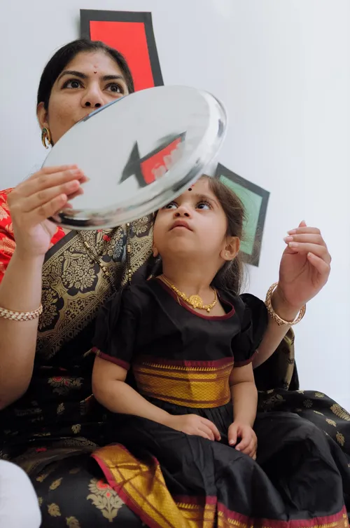A little girl in a traditional black and gold outfit gazes up at a silver plate held above her during a Makar Sankranti Bornhan blessing.