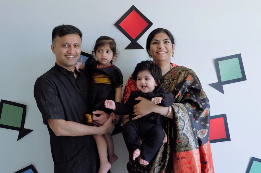 A family of four in festive black outfits stands smiling against a decorated wall, the baby laughing.