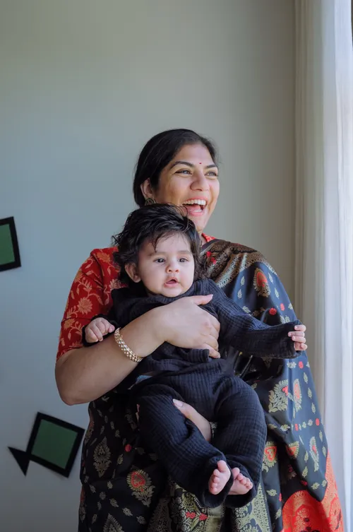 A mother laughs while holding her baby by a sunlit window.