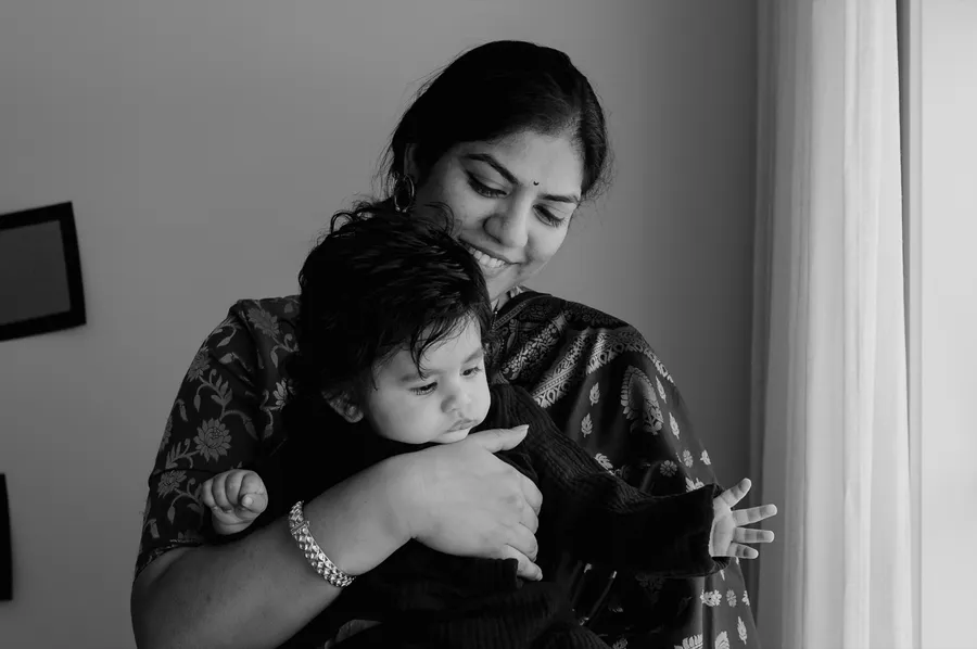 Black and white photo of a mother holding her baby close beside a window.