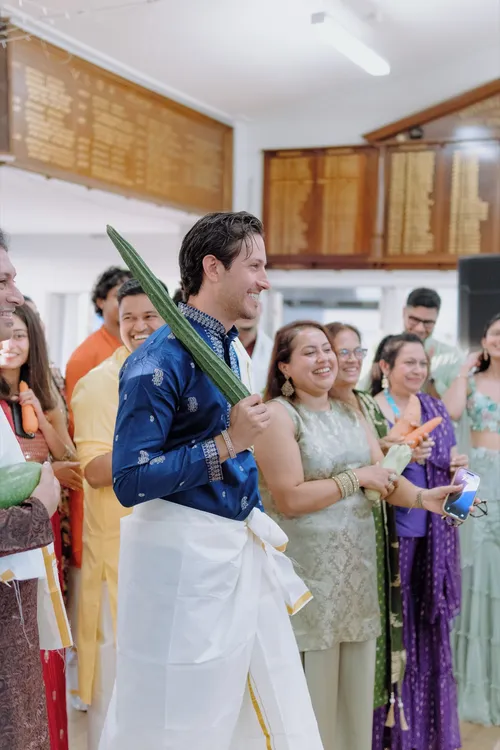 A smiling groom holds a green leaf, surrounded by family in colourful traditional dress at a Roce celebration.