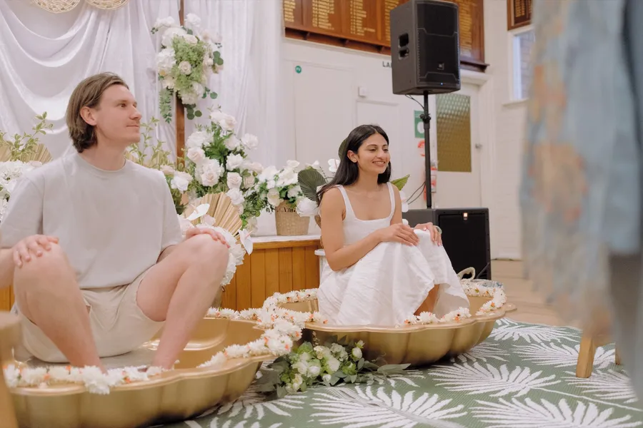 A couple sit with their feet in golden bowls ringed with white flowers during the Roce ritual, the bride smiling.