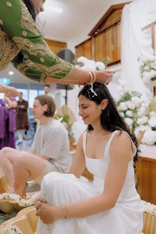 A woman anoints the smiling bride’s hair with coconut milk in front of a white floral backdrop.