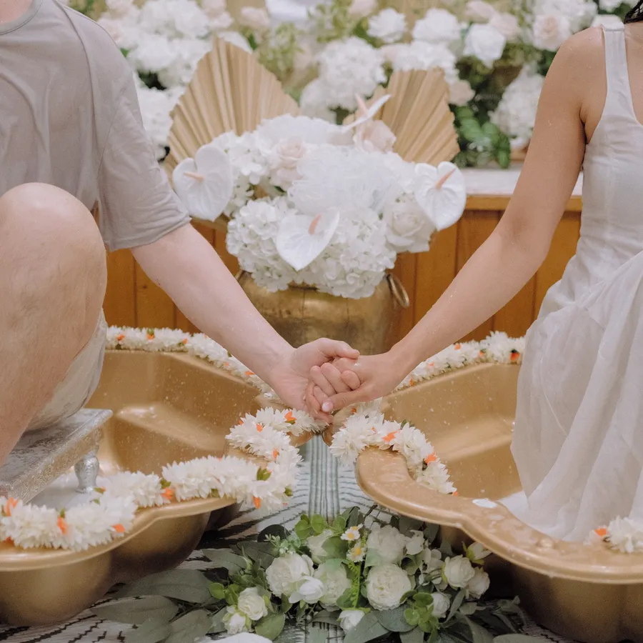 Close-up of the bride and groom holding hands between two golden bowls filled with flowers and coconut milk.