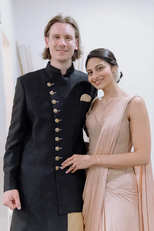 A bride in a rose-gold saree and a groom in a black bandhgala pose together, smiling.