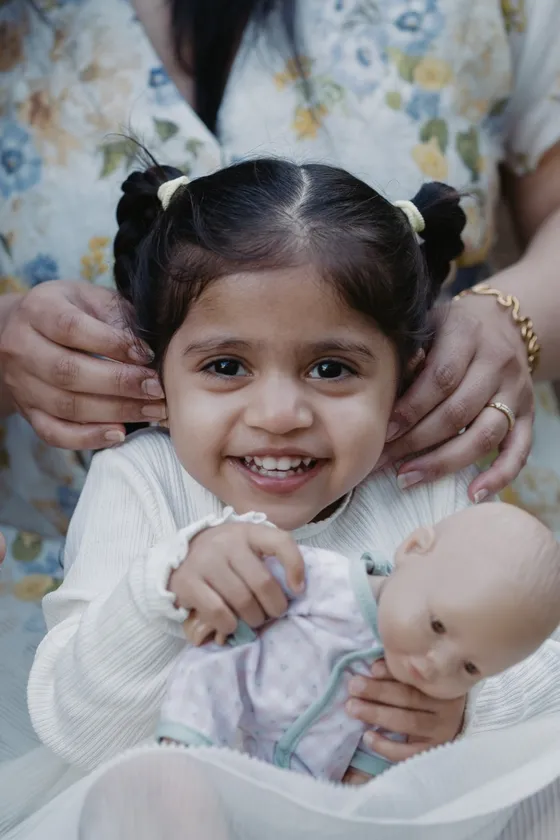 A toddler girl smiles and cuddles her doll while her mum fixes her pigtails.