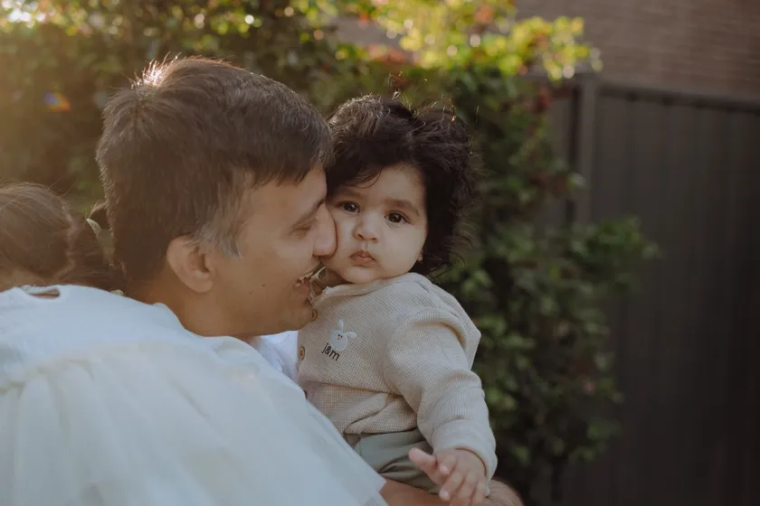 A dad kisses his baby daughter on the cheek in a backyard, late afternoon light behind them.