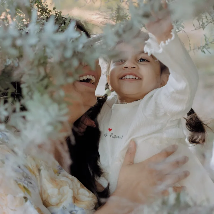 A mum lifts her laughing daughter up through soft wattle branches in dappled light.