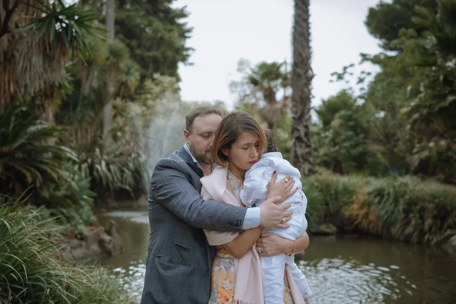 Both parents hold their young son close beside a pond, eyes closed in a quiet, tender moment.