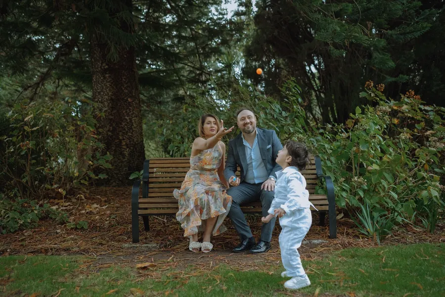 A family plays on a park bench, the dad tossing a small orange as the boy runs in.