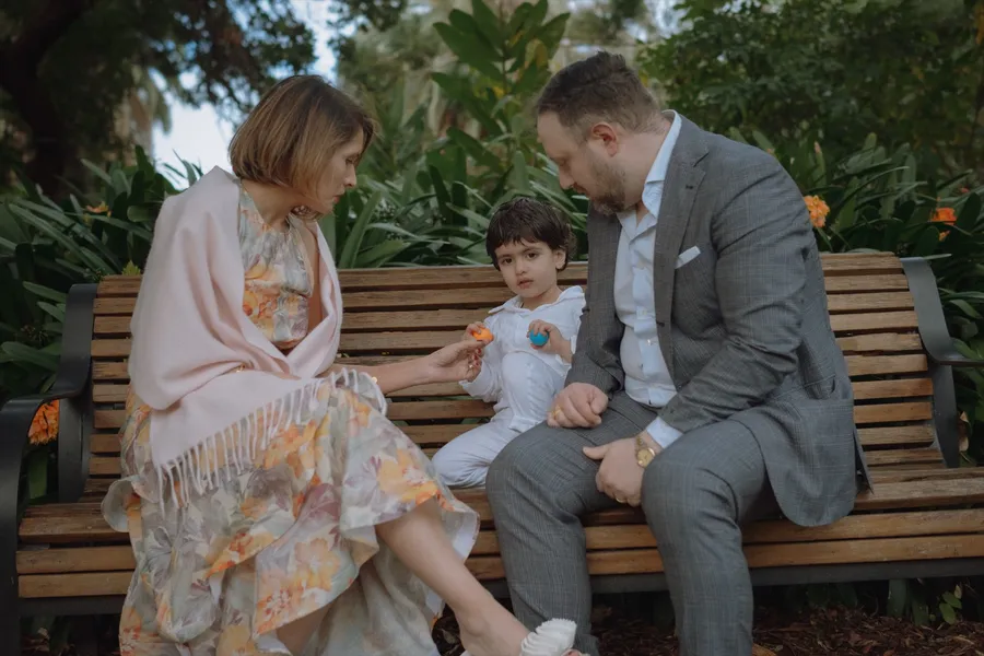 A mother offers something to her son on a park bench while the father looks on, all relaxed.