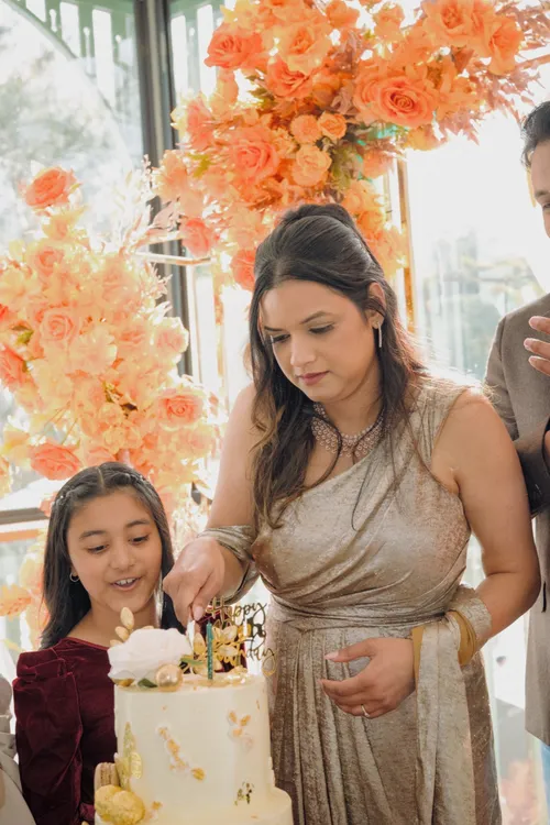 A woman cuts her two-tier birthday cake while her daughter leans in to watch, orange flowers filling the frame behind them.