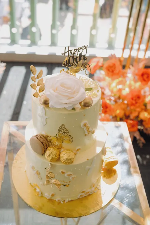 A two-tier white and gold birthday cake topped with a large rose, set against a wall of orange blooms.