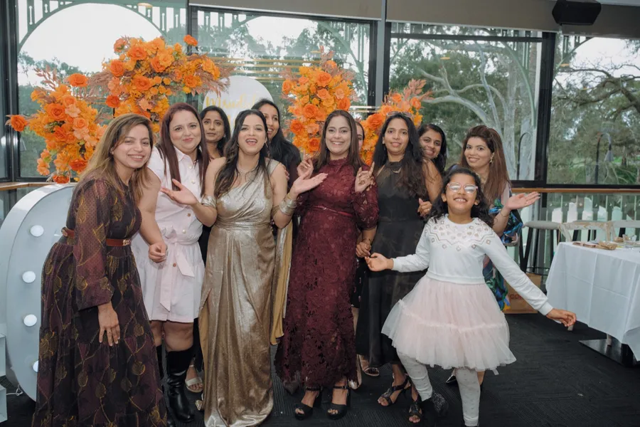 A group of women dance together under an orange floral arch, a small girl in a white dress joining in beside them.
