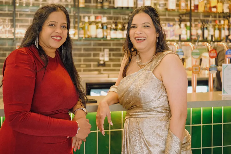 Two guests grin at the camera at the bar, bottles and green tiles behind them.