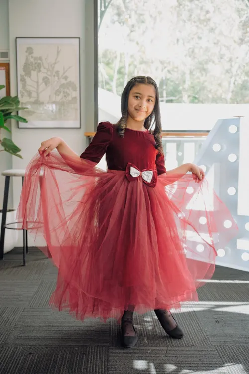 A girl in a deep red tulle dress holds her skirt out wide, smiling.