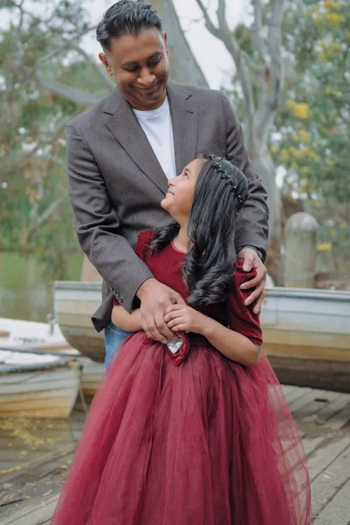A dad hugs his daughter on the boathouse jetty as she looks up at him, rowboats moored behind them.