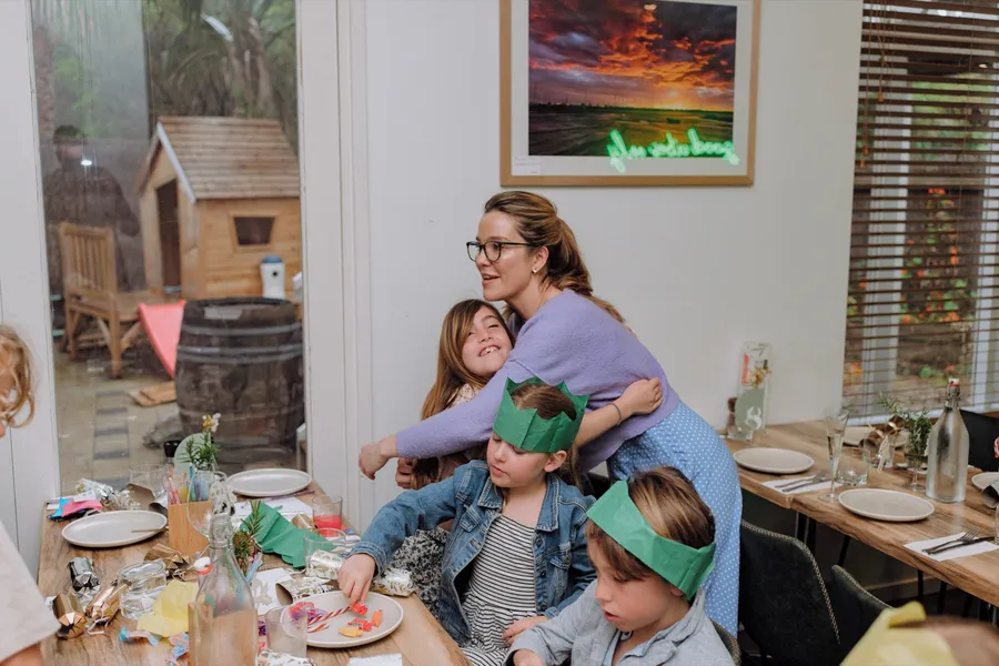A mother hugs her children in green paper crowns around a festive party table.