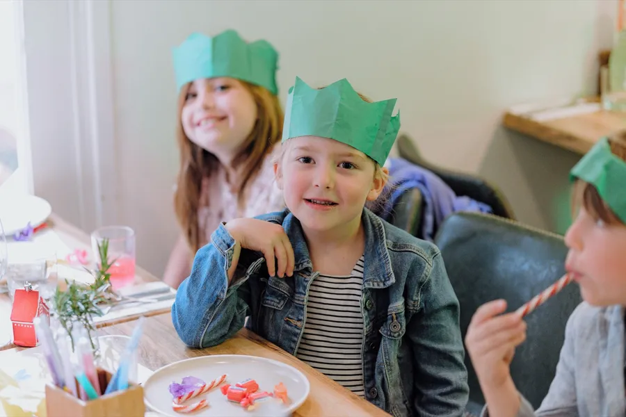 Three children in green paper crowns sit together at a party table, one grinning at the camera and another with a candy cane.