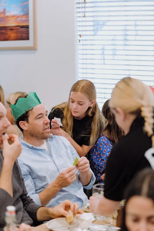 A dad in a green paper crown reads a bon-bon joke while his daughter leans in beside him.