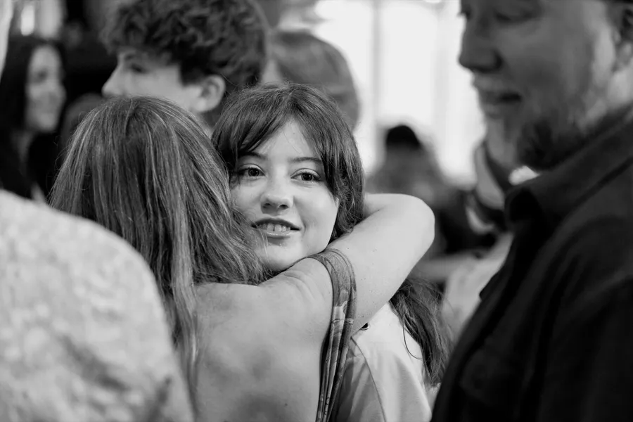 Black and white photo of a young woman smiling over her friend’s shoulder mid-hug at a party.