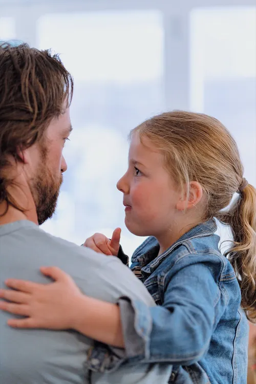 A father holds his young daughter as she talks to him, their faces close together.