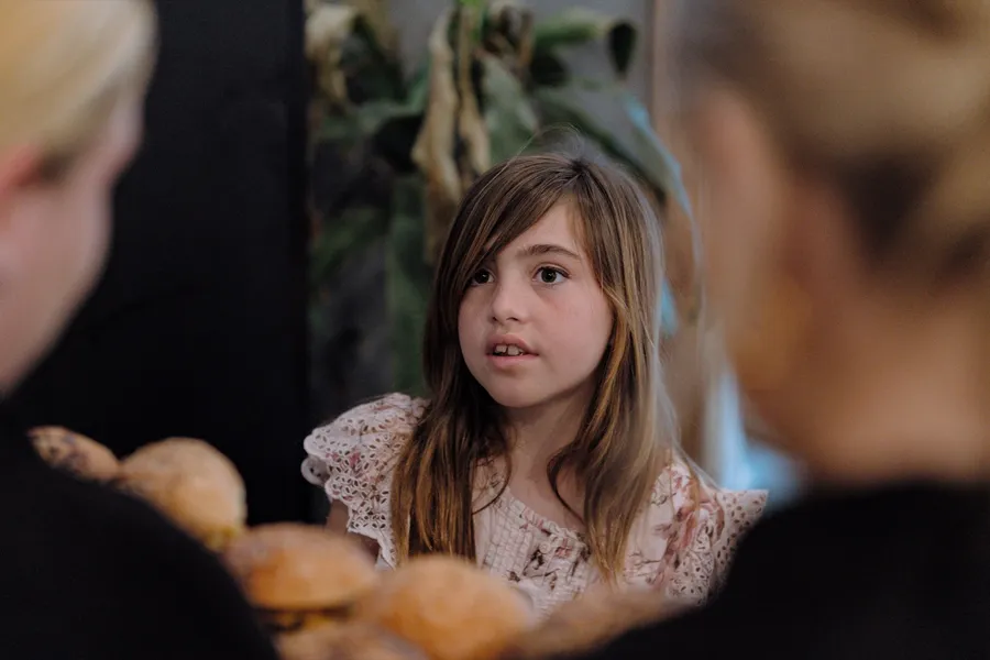 A young girl in a white lace dress chats across the table at a family party.