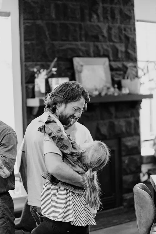 Black and white photo of a dad laughing as he hugs his daughter by a stone fireplace.