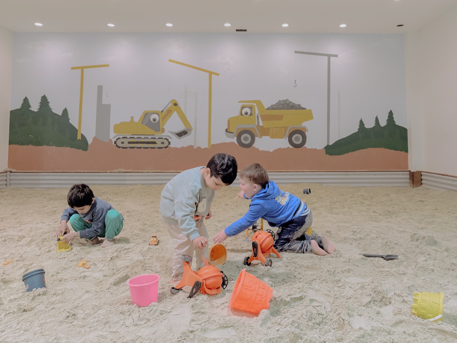Three children play in an indoor construction-themed sandpit, one tipping sand from a toy mixer.