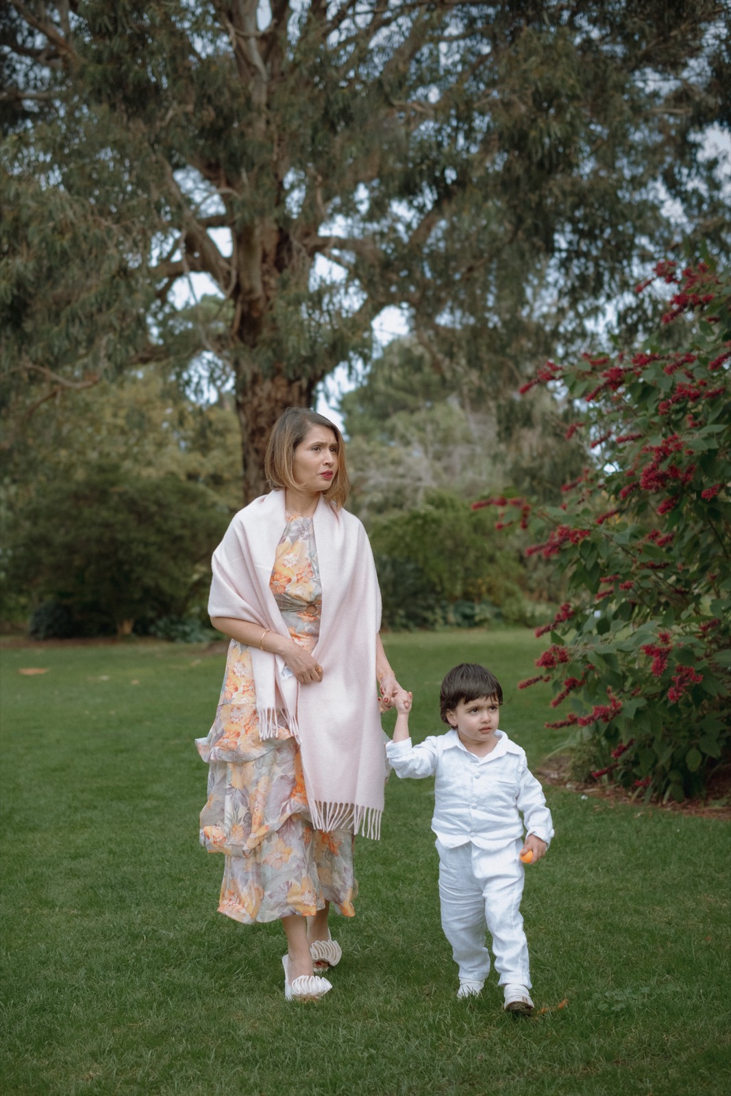 A mother walks hand in hand with her young son past a bush of red berries in the park.