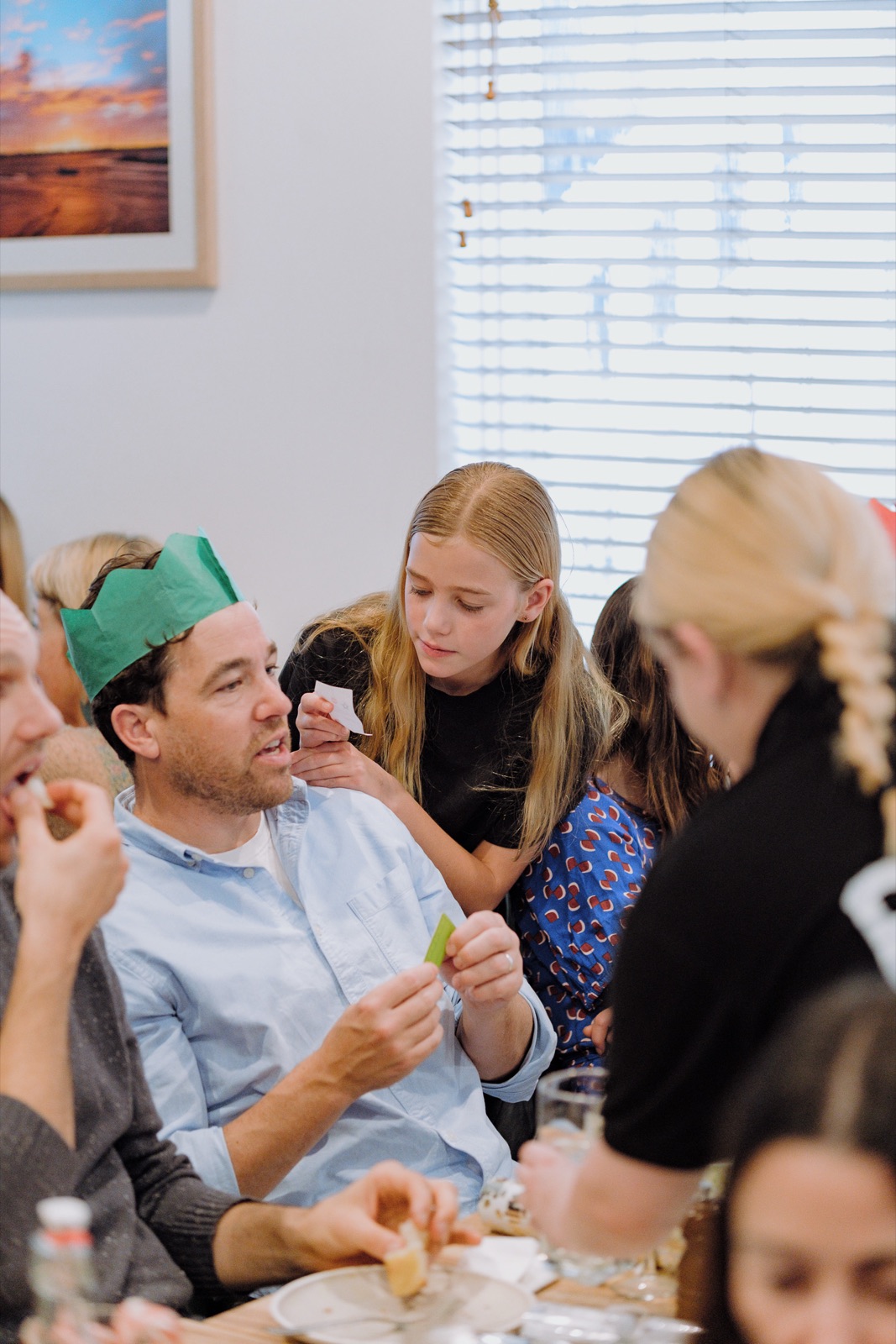 A dad in a green paper crown reads a bon-bon joke while his daughter leans in beside him.